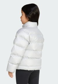 Doudoune en matériau argenté brillant, avec un col haut, un design matelassé et des manches légèrement raccourcies, portée avec des leggings noirs.