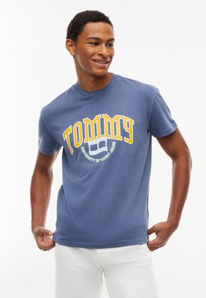 Giovane uomo sorridente, indossa una T-shirt blu Tommy Jeans con testo giallo e pantaloni bianchi, in piedi su uno sfondo bianco uniforme.