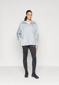 Veste coupe-vent gris clair avec une fermeture éclair à l'avant, capuche ajustable et poche frontale. Leggings de sport noirs avec un motif texturé.