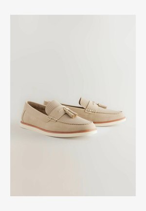 Beige suède instappers met geweven textielaccenten, kwastdetails en een contrasterende bruine loopzool, voorzien van een witte tussenzool voor extra structuur.