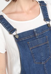 Jeansbyxor med justerbara metallspännen, mörkblå färg, en framficka och avslappnad passform, bärs över en vit topp.