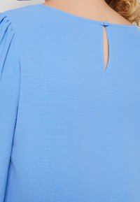 Blusa azzurro chiaro con apertura a occhio di bue sul retro e chiusura con bottone, morbida al tatto e con delicate pieghe sulle spalle.