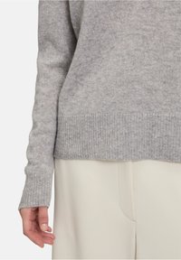 Světle šedý pletený svetr s žebrovaným lemem a manžetami, s jemnou texturou. Spárován s krémovými hladkými kalhotami.