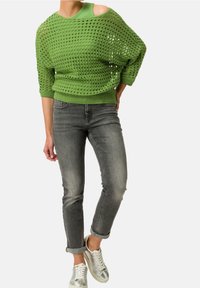 Groene gebreide oversized trui met open schouder, gecombineerd met opgerolde donkergrijze jeans en metallic zilveren sneakers.