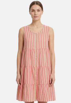 Donna che indossa un vestito beige senza maniche con righe verticali rosa e bottoni frontali, in piedi su sfondo bianco.