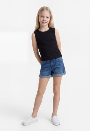 Νεαρό κορίτσι στέκεται με τα χέρια πίσω από την πλάτη, φοράει μαύρο αμάνικο τοπ, τζιν σορτς ρολ, και λευκά παπούτσια slip-on σε λευκό φόντο.