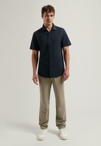 Korteklevig, donkerblauwe button-up shirt van een textuurstof, gecombineerd met lichtbeige broeken en witte sneakers, staande tegen een neutrale achtergrond.