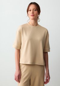 Beżowy top z krótkim rękawem z miękkiego, gładkiego materiału, z okrągłym dekoltem i podwiniętymi rękawami, zestawiony z pasującymi beżowymi spodniami.