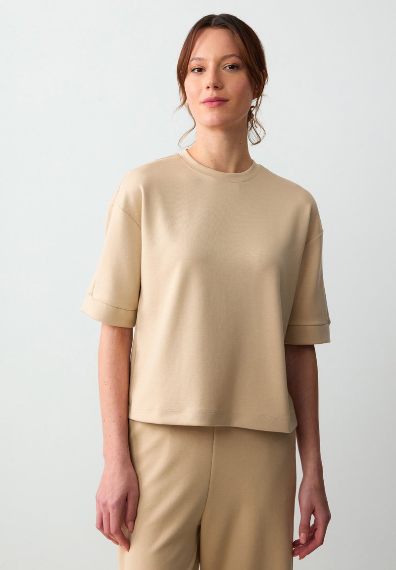 Beżowy top z krótkim rękawem z miękkiego, gładkiego materiału, z okrągłym dekoltem i podwiniętymi rękawami, zestawiony z pasującymi beżowymi spodniami.