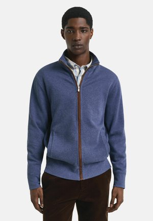 Junger Mann in einer blauen Reißverschlussjacke über einem hellen Hemd mit Kragen und dunklen Hosen, der vor einem schlichten hellen Hintergrund steht.