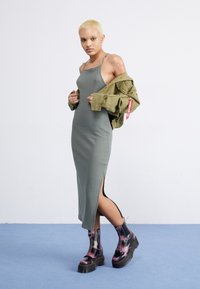 Olivno-zelena rebrasta midi obleka z bočnim razporkom. Model nosi velik olivno-zelen plašč z oznako. Na voljo so črne in roza borbene čevlje.