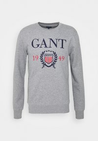 Sudadera gris de cuello redondo, de mangas largas, con el logo "GANT" en azul marino y rojo, y un emblema de escudo con un diseño de laureles.