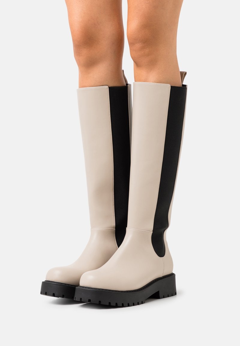 Bottes beige montant jusqu’au genou avec panneaux latéraux élastiques noirs et semelles épaisses noires portées sur jambes nues sur fond blanc.