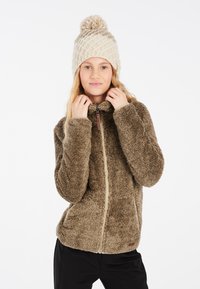 Giacca in pelliccia ecologica marrone con zip frontale, colletto alto e tasche laterali. Indossata con un berretto lavorato a maglia color crema con pompon sopra.