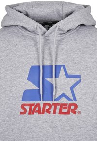 Grå hættetrøje med et stort blåt logo med en stjerne og ordet "STARTER" i rødt. Den har en snor i hætten og et blødt, tekstureret stof.