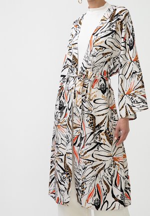 Vrouw draagt een gebloemde kimono-achtige badjas met ceintuur en wijde mouwen over een witte top met hoge hals.