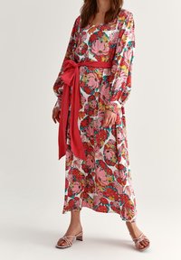 Vestido maxi floral en rosa con flores vibrantes; mangas largas, cintura entallada con una cinta roja, dobladillo fluido, textura de tela suave.