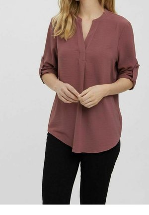 Vrouw die een mauve blouse met lange mouwen en opgerolde mouwen en zwarte broek draagt, met haar handen in haar taille geklemd tegen een effen achtergrond.