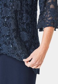 Navyblauwe kanttop met bloemenpatroon, glanzende pailletten en driedelige mouwen, gelaagd over een effen navyblauwe stof aan de onderkant.