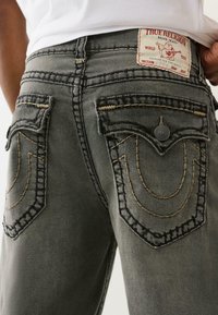 Tylny widok szarych dżinsów True Religion z klapami kieszeni, beżowym przeszyciem i metką marki na pasku, noszonych z białą koszulą.