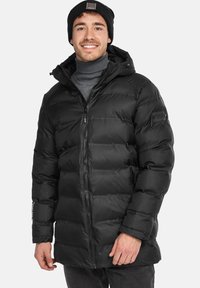 Chaleco negro tipo puffer con capucha, con cierre frontal, bolsillos laterales y un tejido suave y brillante. El modelo lleva un gorro negro a juego.