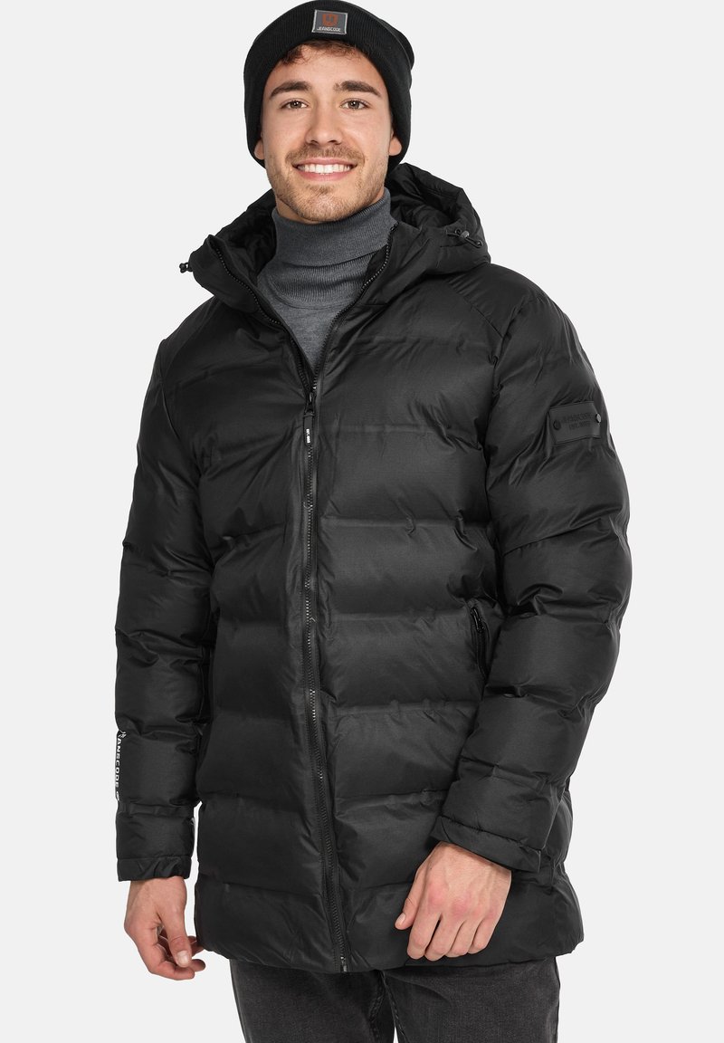Chaleco negro tipo puffer con capucha, con cierre frontal, bolsillos laterales y un tejido suave y brillante. El modelo lleva un gorro negro a juego.