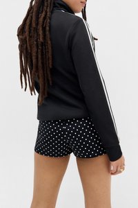 Person mit langen Dreadlocks, der eine schwarze Jacke mit weißen Streifen und schwarze Shorts mit Punkten trägt, steht vor einem einfarbigen Hintergrund.
