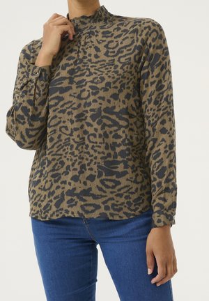 Kvinde iført en langærmet bluse med leopardprint og høj krave samt blå jeans, der står foran en ensfarvet baggrund.