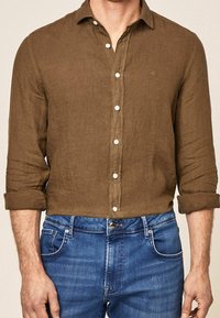 Homme portant une chemise marron à boutons avec manches retroussées et un jean bleu en denim, debout avec les bras détendus sur les côtés.