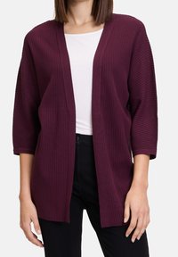 Žena v tmavě fialovém cardigan s vroubkovaným vzorem, otevřeným vpředu a tříčtvrtečními rukávy, přes bílý top a černé kalhoty.