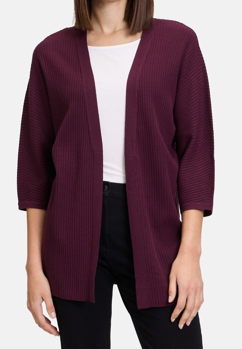 Žena v tmavě fialovém cardigan s vroubkovaným vzorem, otevřeným vpředu a tříčtvrtečními rukávy, přes bílý top a černé kalhoty.