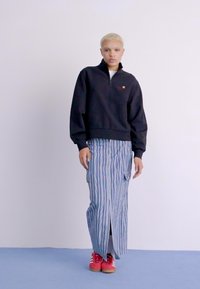 Zwart ritsvest met een logo, gecombineerd met blauw-wit gestreepte wijde broek. Felrode sneakers maken de outfit af.