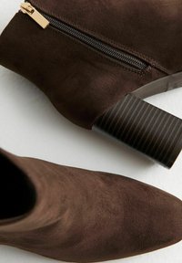 Bruine suede enkelboots met een spitse neus, zijrits met gouden hardware en een gestructureerde zwarte blokhak.