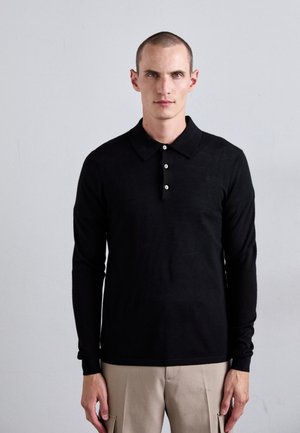 Schwarzes Langarm-Polohemd mit spitzem Kragen und drei weißen Knöpfen; glatte Textur und schlanker Schnitt. Getragen mit beigefarbenen Hosen.
