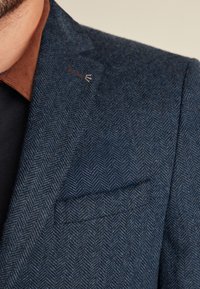 Tweed bleiseri tummansinisellä herringbone-kuviolla, jossa on ruskea corduroy-kaulus ja hopeiset yksityiskohdat läpissä.
