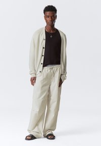 Gilet beige clair avec des boutons noirs, porté sur un t-shirt noir et un pantalon beige clair ample. Des sandales noires complètent le look.