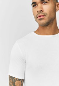 White ribbed short-sleeve T-shirt with a round neckline, shown on a person with visible arm tattoo and neutral background.