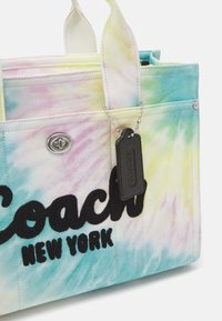 Sac fourre-tout en toile aux couleurs pastel tie-dye avec un texte brodé noir "Coach New York" et une étiquette noire suspendue à une chaîne argentée.