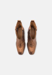 Une paire de bottines métalliques en cuivre avec des motifs décoratifs cousus, des languettes de tirage et des bouts carrés arrondis sur un fond blanc.