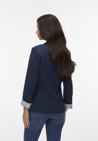 Marineblauw colbert met lange mouwen en gestreepte manchetaccenten, voorzien van een getailleerd ontwerp en een gladde textuur, gecombineerd met een blauwe spijkerbroek.