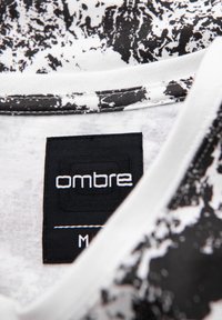 Černobílá textilie s potiskem, která má jemnou strukturu; obsahuje černou etiketu s logem "ombre", bílé prošívání a design s kulatým výstřihem.