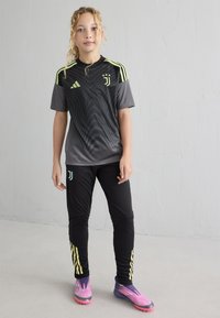 Čierne a sivé športové dresy s horizontálnymi prúžkami, žltými akcentmi a logom. Čierne nohavice so žltými pruhmi a ružovými a fialovými kopačkami.