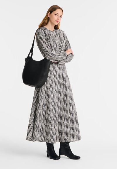 Frau in einem langen gemusterten Kleid, schwarzen Absatzstiefeln und einer großen schwarzen Umhängetasche, die mit verschränkten Armen vor einem einfarbigen Hintergrund steht.