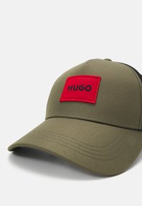 Olivgrön baseballkeps med en röd rektangulär lappar som har "HUGO" i svart, texturerat tyg, och en lätt böjd skärm.