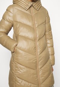 Cappotto puffer beige con motivo a chevron, colletto alto con design a coste e zip frontale. Il materiale ha una finitura lucida.
