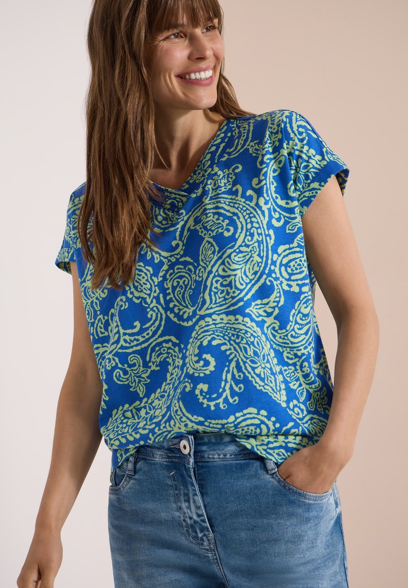 Femme souriante portant un haut bleu à manches courtes avec motif cachemire, rentré dans un jean bleu taille haute, une main dans la poche.