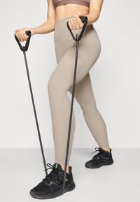 Leggings beige côtelés associés à des chaussures de sport noires, tenant des bandes de résistance en caoutchouc avec des poignées texturées.
