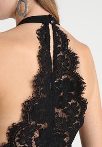 Svart spets halterneck-top med blommönster, ett nyckelhål bak och en knappstängning, med ett mjukt tygband runt nacken.