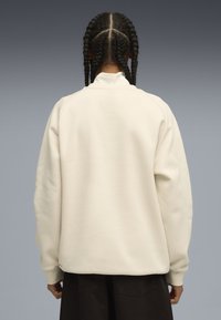 Felpa di colore crema con vestibilità rilassata, polsini a coste e colletto alto, che mostra una texture liscia e morbida senza marchi visibili.