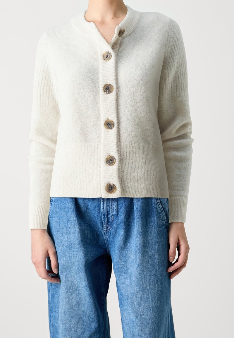 Cream-colored cardigan with a ribbed texture, five round buttons, and a V-neck collar. Paired with blue, loose-fitting denim pants.